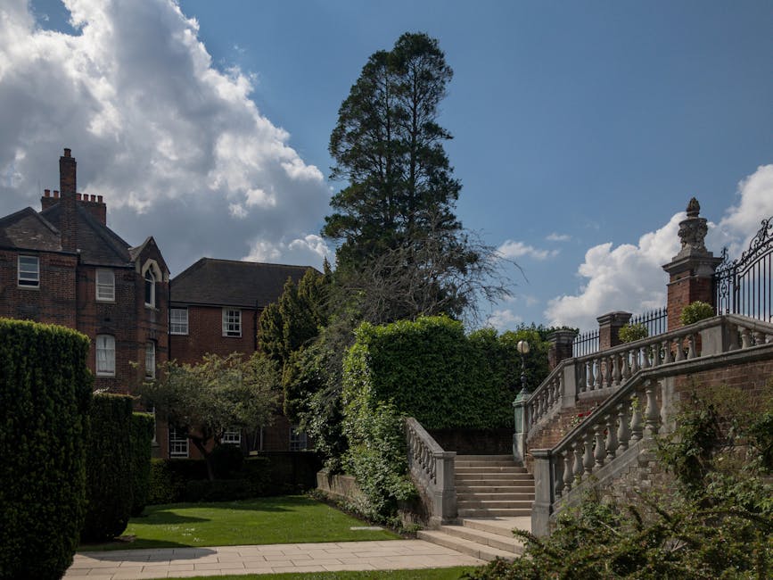 A residential property with a brick exterior and large windows, situated behind a well-maintained garden featuring trimmed hedges, a tall tree, and lush green foliage. The garden area includes a paved pathway leading to an outdoor staircase with stone balustrades, which ascends to an elevated terrace enclosed by decorative iron railings. The staircase is flanked by stone posts and ornate pillars, with a few small plants along the sides. The sky is partly cloudy with patches of blue visible, and sunlight filters through the clouds, casting shadows on the garden and property. This setting appears to be in a suburban neighbourhood, suitable for house removals or relocation logistics, as indicated by the context on manwithvanstanmore.co.uk and the page about access tips for Stanmore removals.
