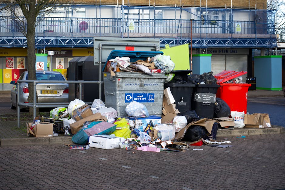 A large outdoor waste collection area with multiple overflowing rubbish bins and bags of waste scattered on the ground. The waste includes cardboard boxes, paper, plastic bags, and packaging materials, some of which are torn or crumpled. The bins are of various colours, including grey, black, and red, with some lids open and waste spilling out. Behind the waste area, there are commercial buildings with shop signs and large windows, and a parked grey vehicle is visible on the left side. The scene is illuminated by natural daylight, and a metal railing separates the waste area from the pavement. This image depicts the disposal of bulky furniture or large waste items, relevant to house removals or furniture transport services that might involve clearing out unwanted items before moving, as offered by Man with Van Stanmore.