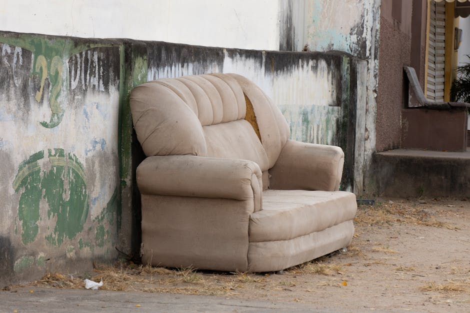 A worn, beige upholstered armchair with visible tears and fabric wear is positioned on a patch of dirt and dried grass, placed against a weathered concrete wall with peeling paint and graffiti in shades of green and white. The chair, part of a home relocation or disposal process handled by Man with Van Stanmore, is situated outdoors near a staircase leading into a residential building, likely awaiting removal or disposal. The surrounding environment includes an urban setting with a concrete sidewalk and an adjacent doorway, indicating the furniture is outside the property, possibly in the process of being loaded onto a van for transport. Nearby, the ground is scattered with small debris and the scene is lit with natural daylight, reflecting a typical furniture removal scenario involving packing up and transporting bulky items during a house move or disposal service.