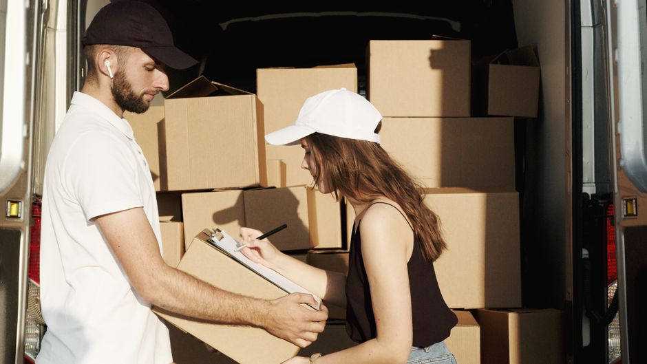 A man and a woman are inside a moving van loaded with stacked cardboard moving boxes, some of which are sealed with packing tape. The man, wearing a white polo shirt, is holding and inspecting a cardb