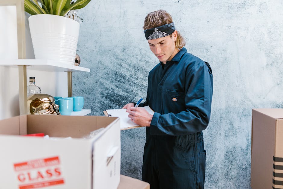 A man and a woman are inside a moving van loaded with stacked cardboard moving boxes, some of which are sealed with packing tape. The man, wearing a white polo shirt, is holding and inspecting a cardb