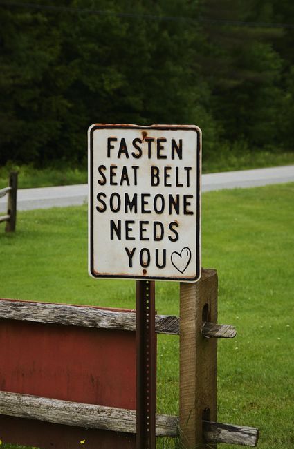 A rectangular metal road sign with a white background and black text reading 'FASTEN SEAT BELT SOMEONE NEEDS YOU' and a small black heart symbol below, mounted on a pole supported by a wooden post. The sign is positioned next to a wooden fence with horizontal slats, adjacent to a grassy area. In the background, there is a rural scene with green trees, a grey road, and a bridge or pathway visible behind the sign. The scene is well-lit, indicating daytime, and the overall setting suggests a peaceful countryside environment. This image can relate to outdoor environments where safety messages are displayed, and while not directly connected to house removals or moving services, it highlights the importance of safety in all transportation contexts managed by professionals like Man with Van Stanmore.