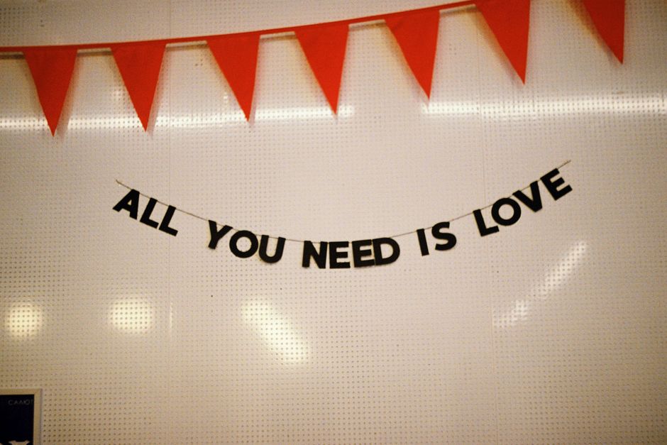 A white interior wall decorated with a black lettered banner that reads 'ALL YOU NEED IS LOVE' suspended from two points. Above the banner, there are multiple red triangular pennant flags strung along the top of the wall. The setting appears to be indoors, with soft lighting reflected on the wall surface. This image may be part of a home or event decoration, potentially during a gathering or celebration. The presence of this decor in a moving or packing context relates to the importance of creating a welcoming atmosphere during a home relocation, as handled by Man with Van Stanmore, which offers removals and transport services. The scene focuses on the decorative display, highlighting a cozy, personable environment.