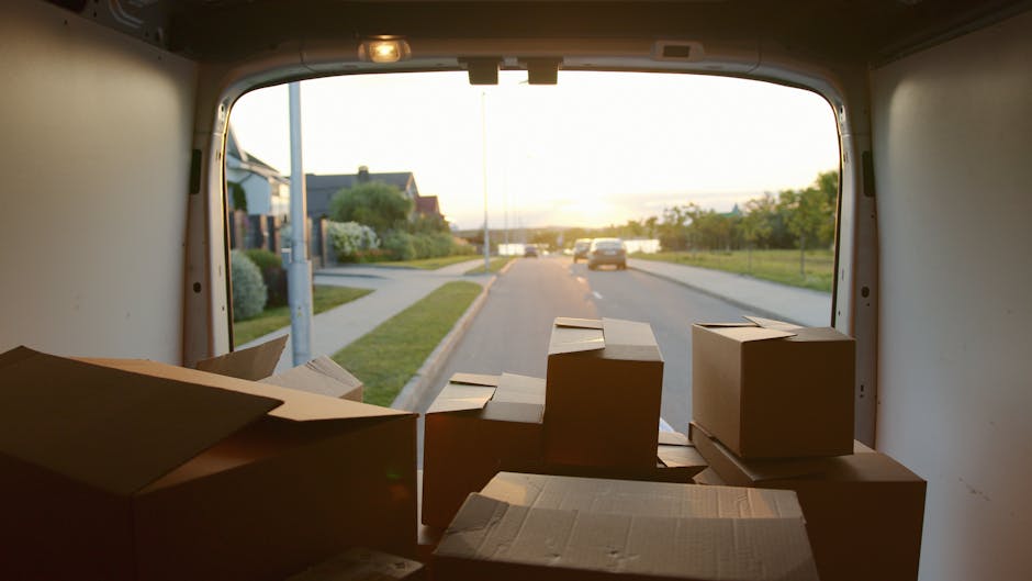 The image shows the interior of a moving van with its rear doors open during daytime, revealing several cardboard boxes of various sizes, some sealed with packing tape and others partially open, stacked on the floor near the doorway. The boxes are arranged towards the back of the van, with a few placed on top of each other on the left side, indicating they are packed for a home relocation. Outside, a residential street is visible with houses, a sidewalk, greenery, and a few parked cars, illuminated by soft natural light from the setting or rising sun. The scene captures the loading process typical for furniture transport and packing during a house removal, with the van positioned on the street preparing to or in the midst of a professional moving service offered by Man with Van Stanmore, supporting efficient and organised relocation logistics.