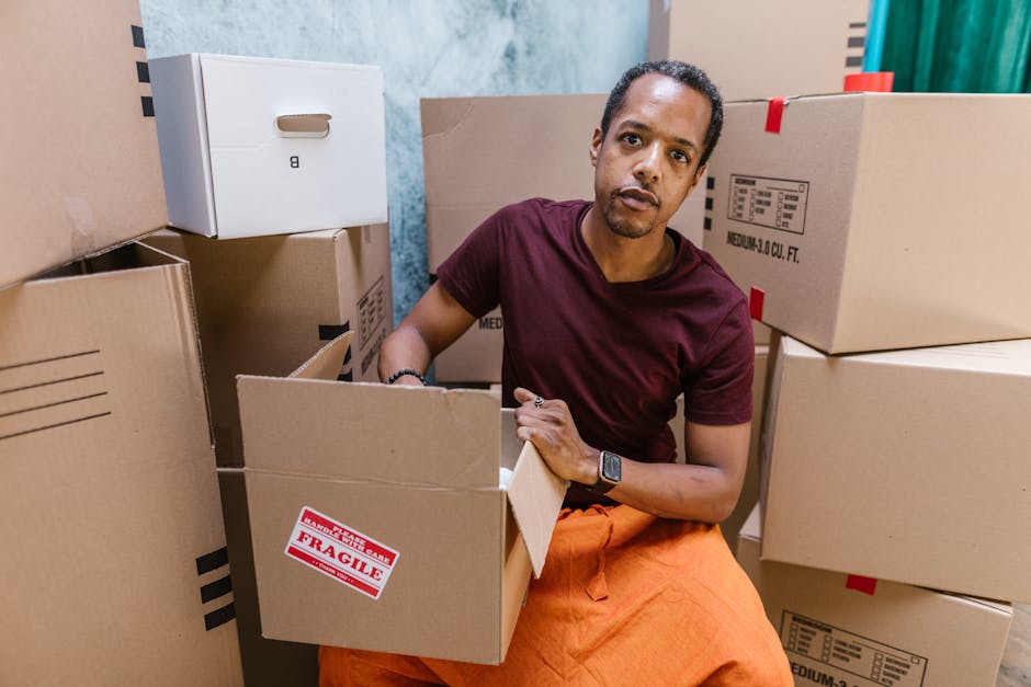 The image shows the interior of a moving van with its rear doors open during daytime, revealing several cardboard boxes of various sizes, some sealed with packing tape and others partially open, stacked on the floor near the doorway. The boxes are arranged towards the back of the van, with a few placed on top of each other on the left side, indicating they are packed for a home relocation. Outside, a residential street is visible with houses, a sidewalk, greenery, and a few parked cars, illuminated by soft natural light from the setting or rising sun. The scene captures the loading process typical for furniture transport and packing during a house removal, with the van positioned on the street preparing to or in the midst of a professional moving service offered by Man with Van Stanmore, supporting efficient and organised relocation logistics.