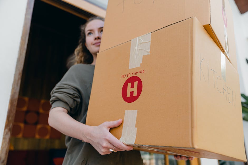 A woman is seen carrying a large cardboard box, which is taped at the top and has the word 'KITCHEN' handwritten on its side. She is inside a home, near a doorway, with the box held securely against her body using both hands. The box features printed red and white markings, including a large red 'H' symbol, and is likely part of a packing process for a home relocation. The background shows a glimpse into a room with a wooden door frame and shelves containing wooden cylinders, indicating the moving is taking place inside a house. The scene is illuminated by natural indoor lighting, capturing the careful handling of packed belongings as part of furniture transport and packing during a house removal. The image reflects a sequence of packing and loading activities associated with professional removals, with the woman preparing items for transport by [COMPANY_NAME], which specializes in move logistics and house removals.