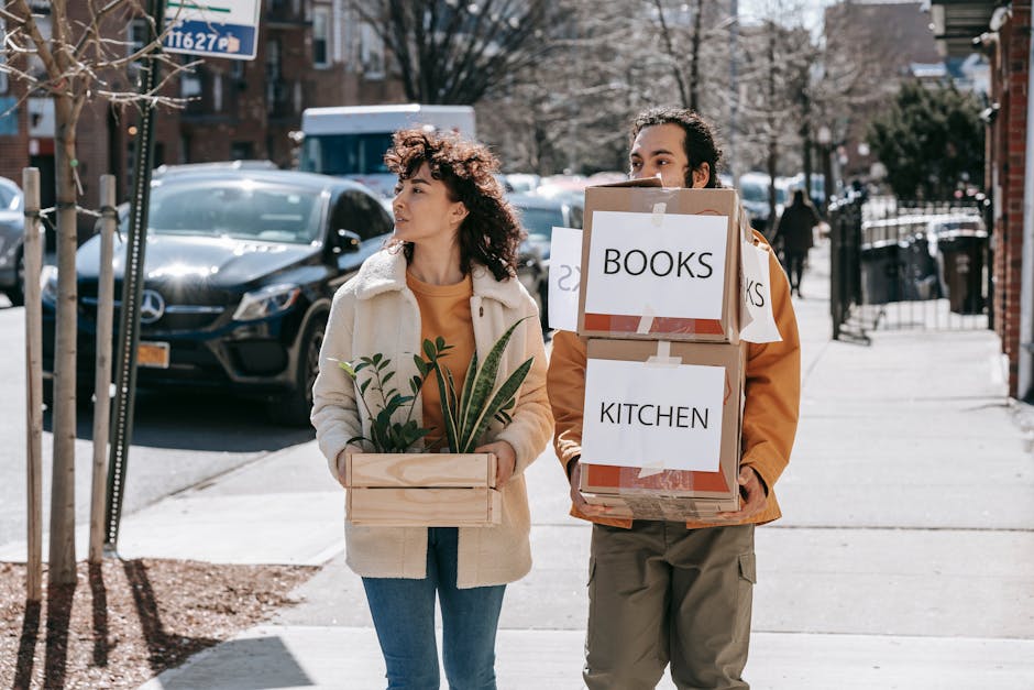 A man and a woman are walking along a residential sidewalk during daytime, each carrying cardboard boxes filled with household items. The woman, on the left, is holding a rectangular wooden crate containing potted plants with green foliage and a small yellow flower, and she is wearing a light-colored fleece jacket over a brown top, with curly hair. She is looking to her right. The man, on the right, is carrying two cardboard boxes stacked on top of each other, labeled 'BOOKS' and 'KITCHEN,' with packing tape securing their flaps. The man is dressed in a brown jacket and beige pants, with dark curly hair, and is looking ahead. In the background are parked cars on the street, including a black vehicle, trees with branches, and residential buildings with brick facades and front gardens. The scene is lit by natural daylight, depicting a home relocation process involving packing and moving furniture and household items, with the presence of a moving company, Man with Van Stanmore, ensuring secure and efficient furniture transport.
