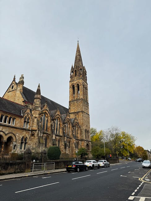 A view of a historic stone church with a tall, pointed spire situated on a street corner, adjacent to a paved road with parked cars, including a black sedan, white vehicles, and a grey car, in a resid