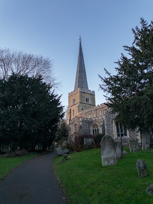 A view of a historic stone church with a tall, pointed spire situated on a street corner, adjacent to a paved road with parked cars, including a black sedan, white vehicles, and a grey car, in a resid