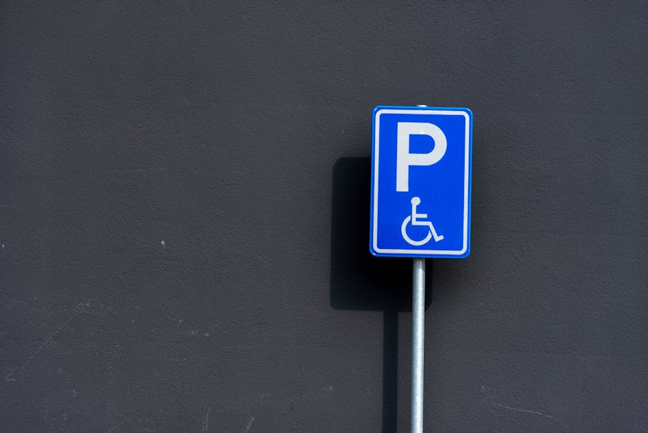 A blue parking sign with white symbols indicating accessible parking is mounted on a metal post in front of a dark grey textured wall. The sign displays a large white letter 'P' and a wheelchair symbol below it, designating designated disabled parking. The shadow of the sign is cast onto the wall, suggesting a light source from the left side of the image. The scene is outdoors, with no vehicles or other objects visible around the sign. This setting relates to parking regulations often encountered during house removals or furniture transport, and the sign’s placement indicates available accessible parking space likely relevant for relocating services by Man with Van Stanmore as part of their moving logistics. The image emphasizes the importance of planning around parking restrictions during home relocation tasks, especially in urban areas like Stanmore, where permits may be required from Harrow Council.