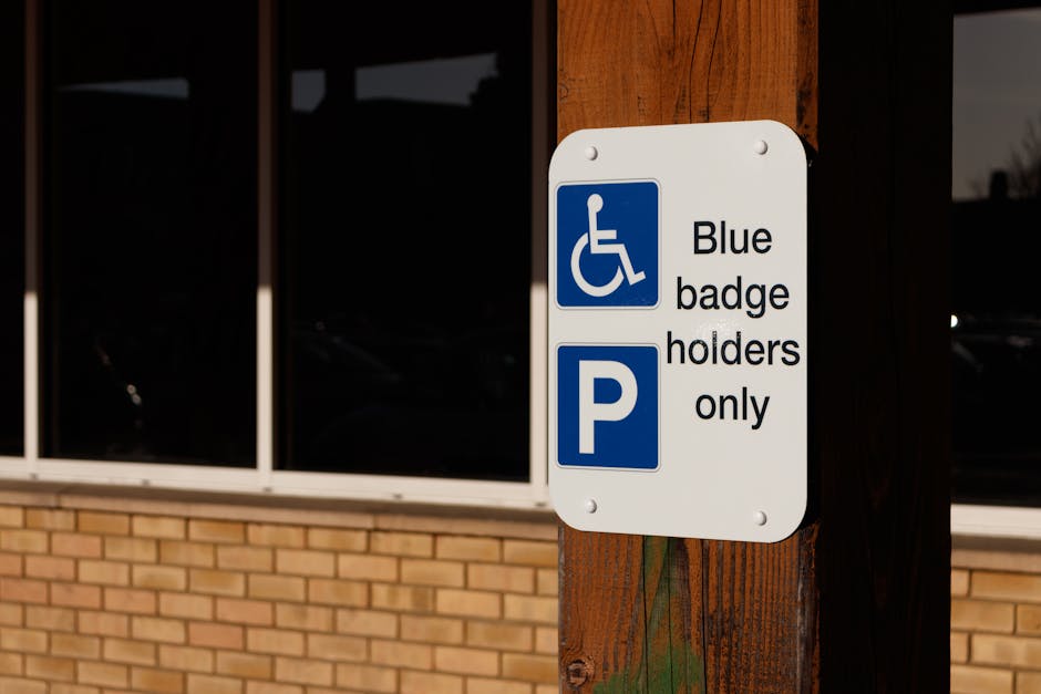 A close-up photograph of a white rectangular parking sign attached to a wooden post outside a building with large glass windows and brickwork. The sign features a blue wheelchair accessibility symbol above the text 'Blue badge holders only' and a blue 'P' symbol indicating parking. The background shows the dark reflections of parked cars in the windows, and the scene is illuminated by natural daylight. This image relates to parking regulations in the context of house removals or relocation services provided by Man with Van Stanmore, highlighting accessible parking provisions in the area near the property for ease of furniture transport and home relocation logistics.