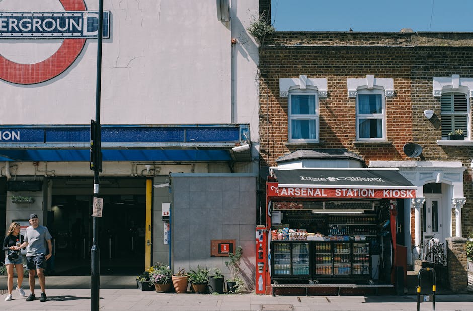 The image shows the exterior of Hammersmith underground station in London, with train tracks running through the station platform. The station features a covered waiting area with a curved, translucen