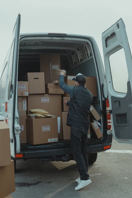 A person wearing a black jacket, black pants, white sneakers, and a black cap is standing next to an open white delivery van parked outdoors on a paved surface. Inside the van, there are multiple cardboard boxes of various sizes, some labeled with symbols indicating fragile contents, and a few padded fabric or plastic-wrapped items. The individual is in the process of loading or organizing boxes, with their right hand reaching towards one of the boxes. The van's interior appears spacious, with a high roof and storage shelves, and the doors are fully open to facilitate loading. Surrounding the vehicle is an outdoor scene with natural lighting, and the background shows an overcast sky. This scene illustrates a home relocation or professional moving process conducted by Man with Van Stanmore, focusing on furniture and packing logistics for house removals or shorter-distance moves, including the use of appropriate packaging materials and loading procedures.