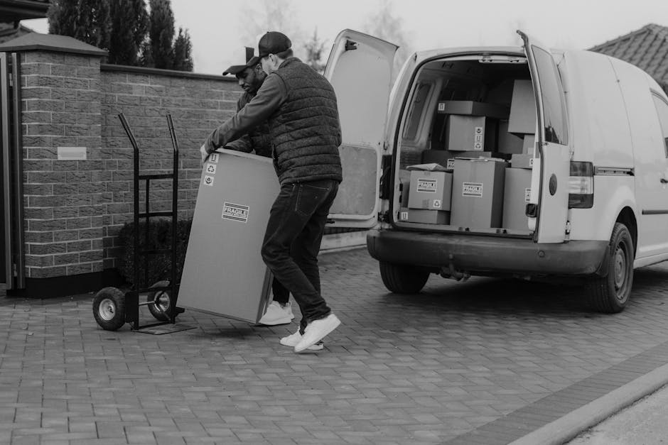 Two men engaged in a home relocation process are seen loading a cardboard box onto a hand truck outside a residential property. One man is wearing a cap, dark jacket, and jeans, while the other is dressed in a hoodie and sneakers. They are positioned on a paved driveway next to a white van with its rear doors open, revealing several packed boxes inside. The boxes vary in size and are made of cardboard, some sealed with packing tape. A brick wall and a garden area are visible in the background. The scene captures the loading process involved in furniture transport and packing during a house move, with the men carefully handling the boxes to ensure safe transport, illustrating the logistics managed by [COMPANY_NAME] for quick and efficient relocation services in Stanmore.