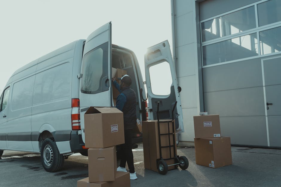 A person wearing a black jacket, black pants, white sneakers, and a black cap is standing next to an open white delivery van parked outdoors on a paved surface. Inside the van, there are multiple cardboard boxes of various sizes, some labeled with symbols indicating fragile contents, and a few padded fabric or plastic-wrapped items. The individual is in the process of loading or organizing boxes, with their right hand reaching towards one of the boxes. The van's interior appears spacious, with a high roof and storage shelves, and the doors are fully open to facilitate loading. Surrounding the vehicle is an outdoor scene with natural lighting, and the background shows an overcast sky. This scene illustrates a home relocation or professional moving process conducted by Man with Van Stanmore, focusing on furniture and packing logistics for house removals or shorter-distance moves, including the use of appropriate packaging materials and loading procedures.