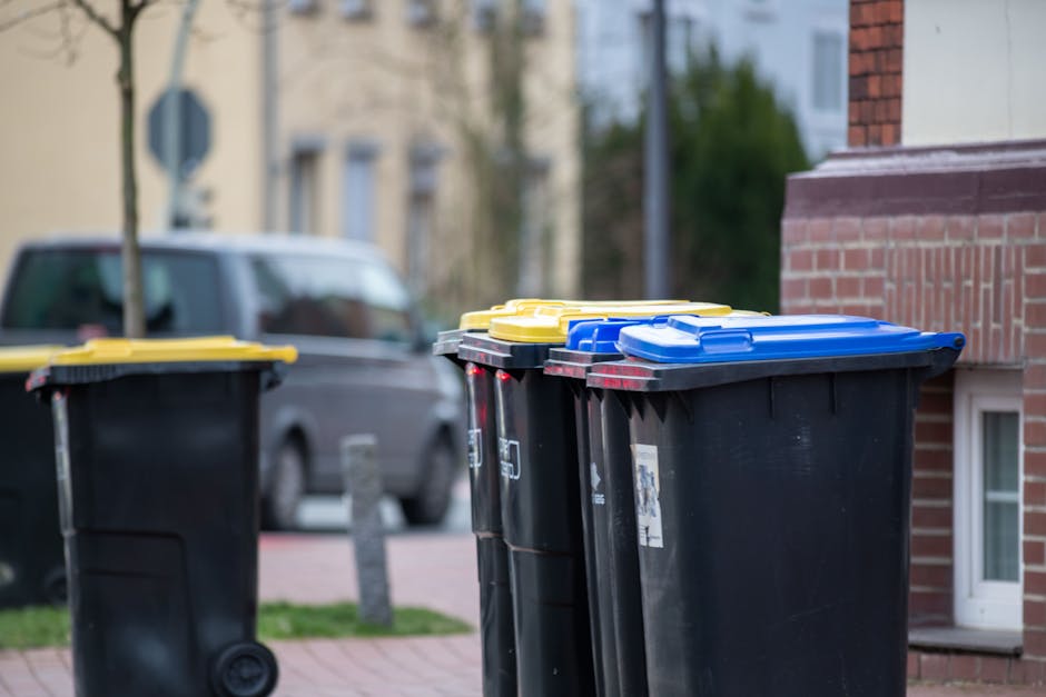 A row of four large wheeled bins with yellow, blue, and black lids, positioned on a pavement outside a residential building. The bins are intended for waste disposal and recycling, with some lids partially open. Behind the bins, a parked grey van is visible on the street, with residential buildings featuring brick and stucco facades in the background. The environment appears to be in daylight with natural lighting. This scene illustrates common waste collection arrangements near homes, relevant to house removals and the proper disposal process as part of a home relocation or packing and moving tasks, which Man with Van Stanmore occasionally handles in their removals services.