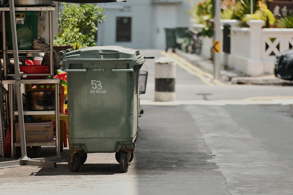 A green wheeled waste bin with the number 53 and the words 'JUN BESAR' printed on the side is positioned on the sidewalk outside a residential property, close to a storage shelf filled with various household items, including plastic containers and packaged goods. The bin is situated near the entrance to a driveway, with a paved street visible in the background, along with a bollard, a traffic mirror, and some greenery in the distance. The scene is illuminated by natural daylight, illustrating a typical outdoor setting where waste disposal or clearance activities might be occurring during a home removal or moving process, as handled by Man with Van Stanmore, ensuring proper waste disposal compliance during relocation.