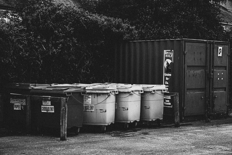A row of four large wheeled bins with yellow, blue, and black lids, positioned on a pavement outside a residential building. The bins are intended for waste disposal and recycling, with some lids partially open. Behind the bins, a parked grey van is visible on the street, with residential buildings featuring brick and stucco facades in the background. The environment appears to be in daylight with natural lighting. This scene illustrates common waste collection arrangements near homes, relevant to house removals and the proper disposal process as part of a home relocation or packing and moving tasks, which Man with Van Stanmore occasionally handles in their removals services.
