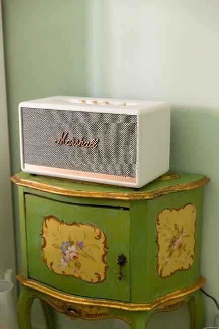 A white Marshall speaker is placed on top of a vintage green wooden cabinet with curved edges and floral painted details, positioned against a green wall. The cabinet features two small doors decorated with painted floral motifs in yellow and pink, with a black metal handle and a decorative keyhole. The scene is inside a home, with natural lighting illuminating the objects, suggesting a tidy indoor environment. This setup may be part of a moving or packing process, where fragile electronics are being prepared for transportation, and the cabinet might be temporarily used as a staging surface. Man with Van Stanmore offers house removals and furniture transport services, including careful handling of delicate items during home relocations such as this.