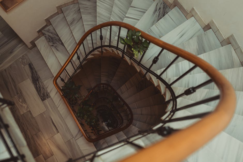 The image shows a staircase viewed from inside a building or parking area, with dark walls and a concrete floor. The stairs are made of concrete or stone and ascend towards an outdoor area with terraced steps. At the top of the stairs, there are residential buildings with brick walls, metal railings, and potted plants with green foliage along the pathway. The scene is illuminated by natural daylight, casting shadows on the steps and surrounding surfaces. This setting reflects the typical environment involved in house removals and furniture transport, with the staircase presenting a narrow, potentially challenging passage for a home relocation or moving process. A professional moving service like Man with Van Stanmore would carefully manage such spaces during their packing and loading operations, ensuring furniture and boxes are safely navigated through stairways during the lifting and transport phases of a home removal.